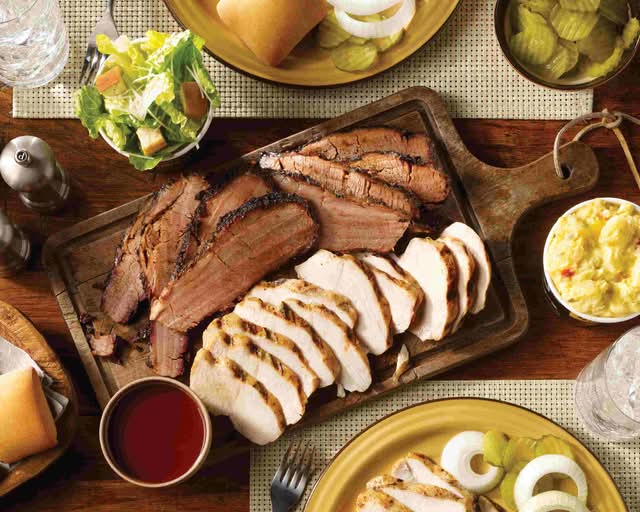 An overhead shot of a large platter with sliced brisket and turkey, served with salads, rolls, and pickles.